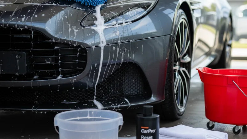 Auto wassen zonder fouten thuis: emmer met zeepwater en microvezel washandschoen naast auto op oprit